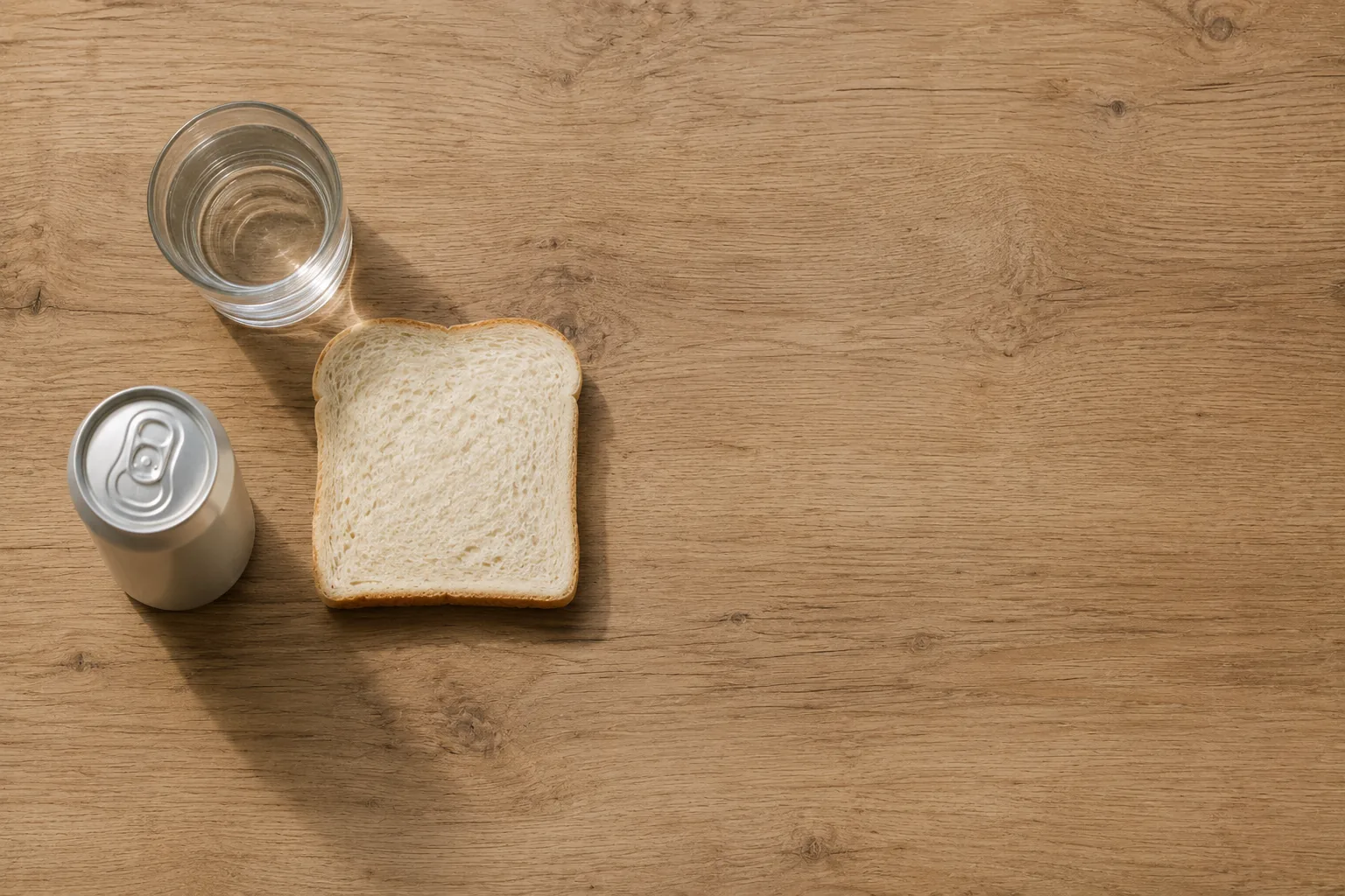 A slice of white bread, a can of soda and a glass of water on a plain wooden table, shot from above.