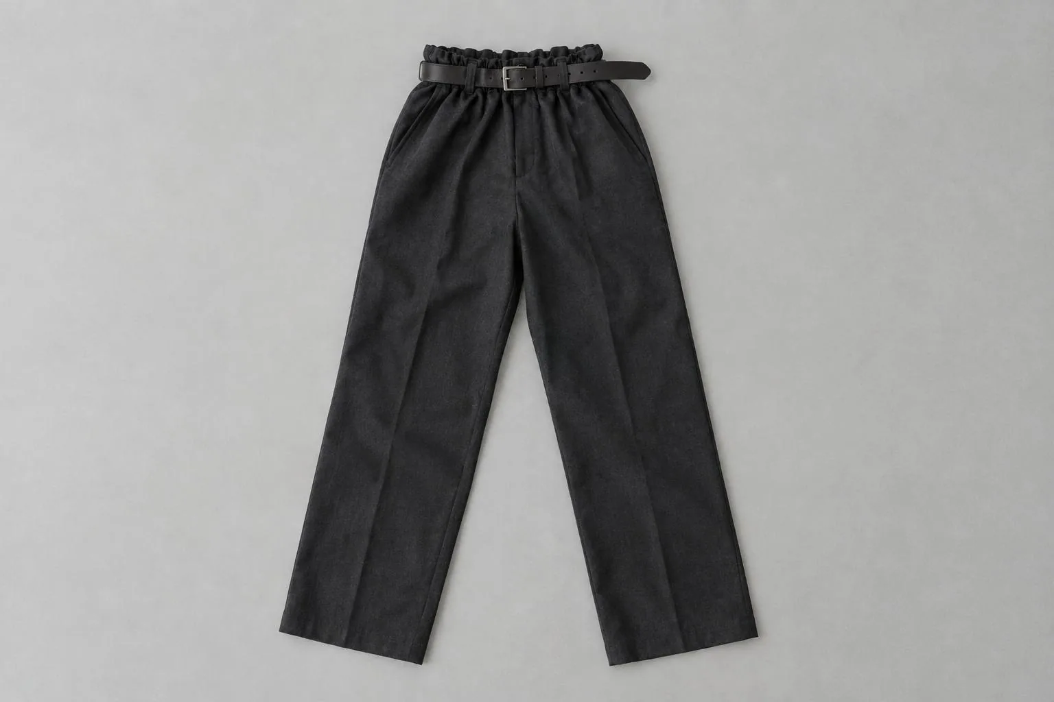 A pair of dark charcoal trousers laid flat, with a leather belt pulled tight so the fabric gathers and bunches at the waist.